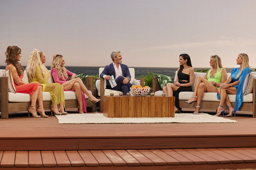 Emily Simpson, Shannon Storms Beador, Tamra Judge, Andy Cohen, Heather Dubrow, Gina Kirschenheiter, and Jennifer Pedranti seated in front of a sunset.