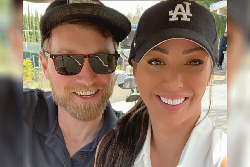 Kristen Doute and Luke Broderick wearing baseball caps and polo shirts as they take a selfie.