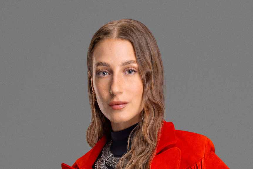 Amanda Batula in a red, fringed, blazer, a black turtleneck, and a silver statement necklace in front of a grey backdrop.