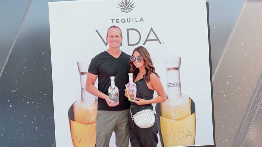 Lisa Barlow and John Barlow holding bottles of Vida tequila in front of a large Vida tequila poster.