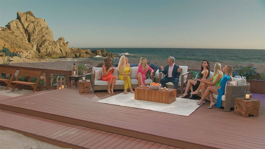 Andy Cohen and the RHOC cast sit on the reunion stage in front of a sunset, seaside, background.