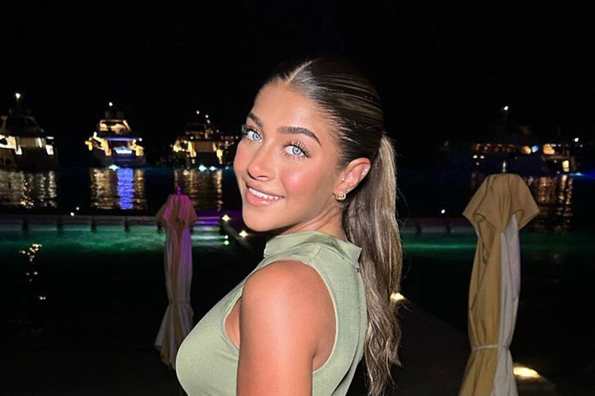 Gia Giudice posing in an olive green, 2 piece, outfit in front of a beach and waterfront at night.