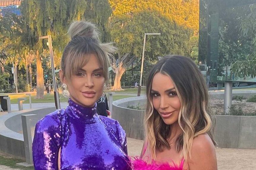 Lala Kent and Scheana Shay pose in pink and purple cocktail dresses in a driveway.