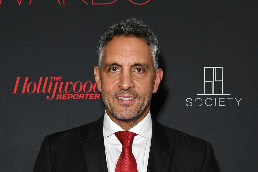 Mauricio Umansky smiling in front of a step and repeat wearing a black suit, white button down, and red tie.