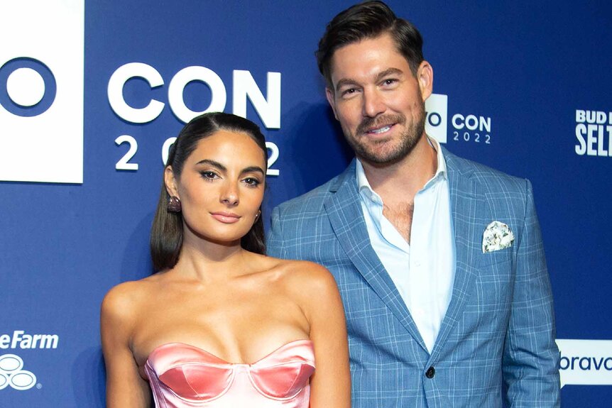 Paige DeSorbo wears a pink strapless dress with Craig Conover, who wears a blue suit, in front of a BravoCon 2022 step and repeat.