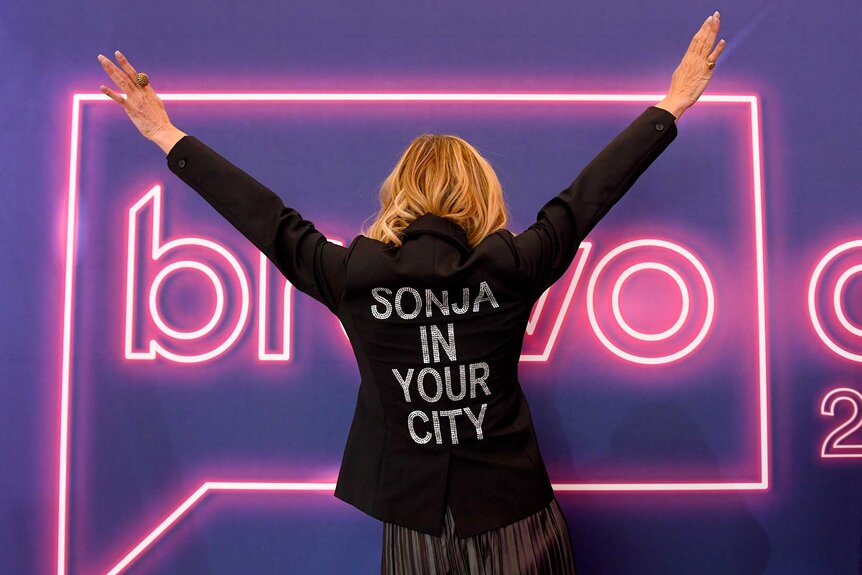 Sonja Morgan wearing a jacket that says "Sonja In Your City" with both her hands raised at BravoCon 2023.
