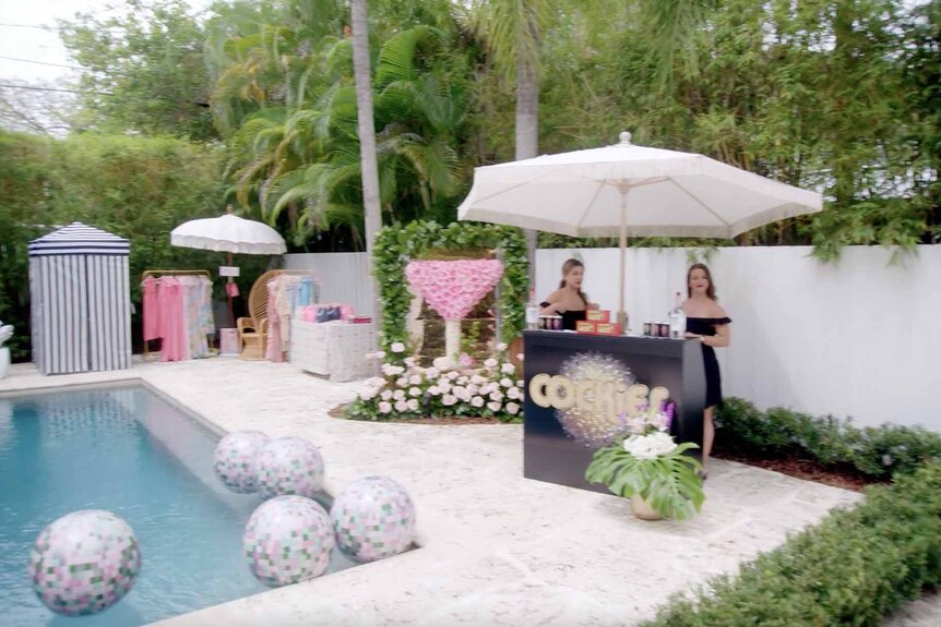 Two female bartenders stand under an umbrella stand near a pool at Marysol Patton's party.