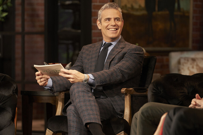 Andy Cohen smiling while holding cue cards.