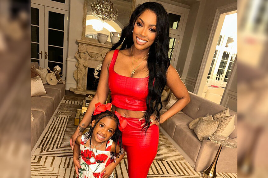 Porsha Williams wearing a red top and pants with her daughter Pilar Jhena in a living room.
