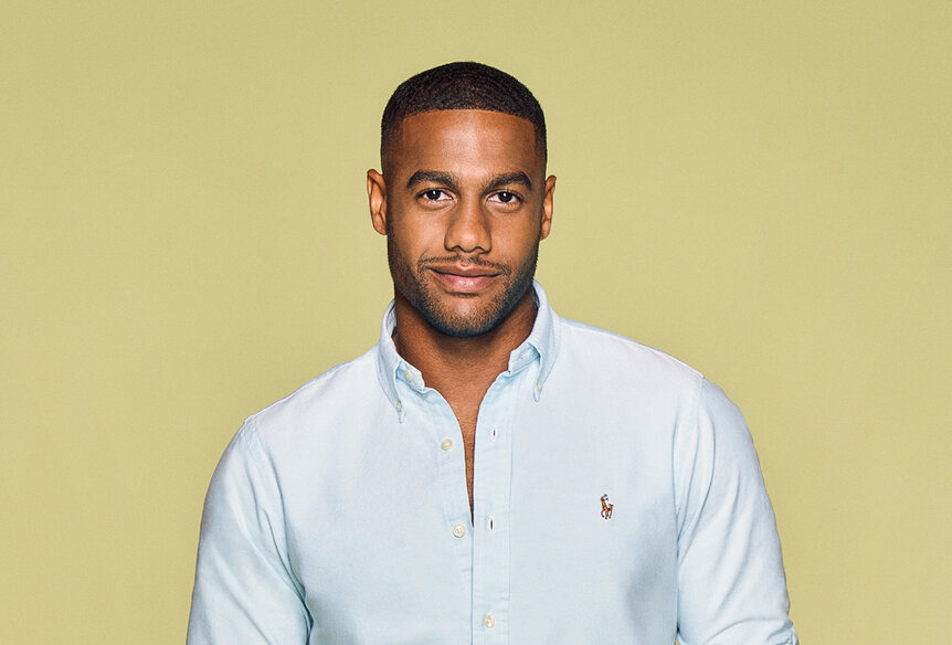 Amir Lancaster wearing a light blue button down in front of a light brown backdrop.